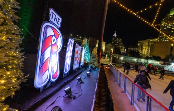 Outdoor holiday ice skating rink at night in a city center, decorated with Christmas lights and illuminated signage reading “The Rink” in festive candy cane and gift box designs. Skaters glide on the ice while surrounding Christmas trees with twinkling lights add to the festive atmosphere. Warm string lights hang overhead, and tall city buildings glow in the background, creating a lively winter holiday scene.